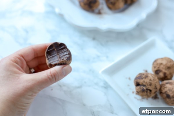 5 Malzemeli Baştan Çıkarıcı Vegan Trüf 2 A hand delicately holds a half-eaten vegan truffle, revealing its rich, dark chocolate interior. In the background, plates laden with more perfectly rolled truffles suggest a bountiful dessert spread, inviting indulgence.