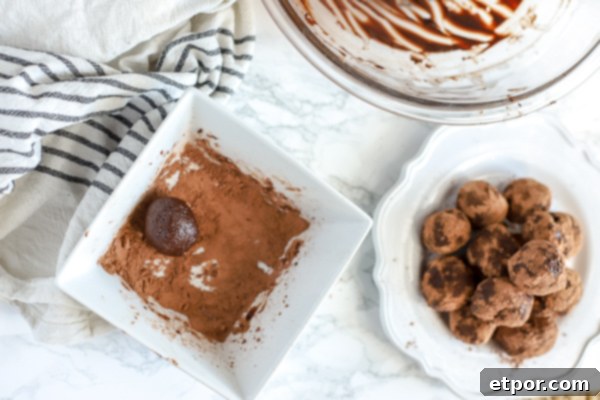 5 Malzemeli Baştan Çıkarıcı Vegan Trüf 5 A selection of freshly rolled vegan truffles being coated in a bed of cocoa powder, with a pristine stack of finished truffles on a white plate to the right. The remaining ganache is visible in the background, all set against a subtly striped kitchen towel to the left, capturing the essence of a rewarding baking process.