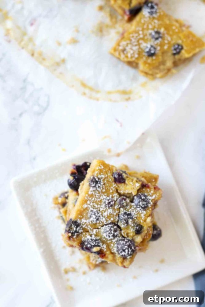 blueberry lemon bars on a white plate and dusted with powder sugar. More sliced bars on the right on top of parchment paper