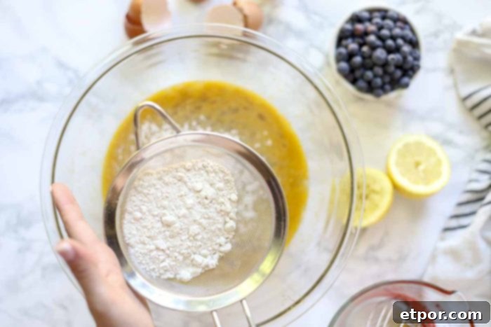 hand holding a fine mesh strainer with flower over a bowl of eggs, lemon juice, sugar. Other ingredients surround the bowl
