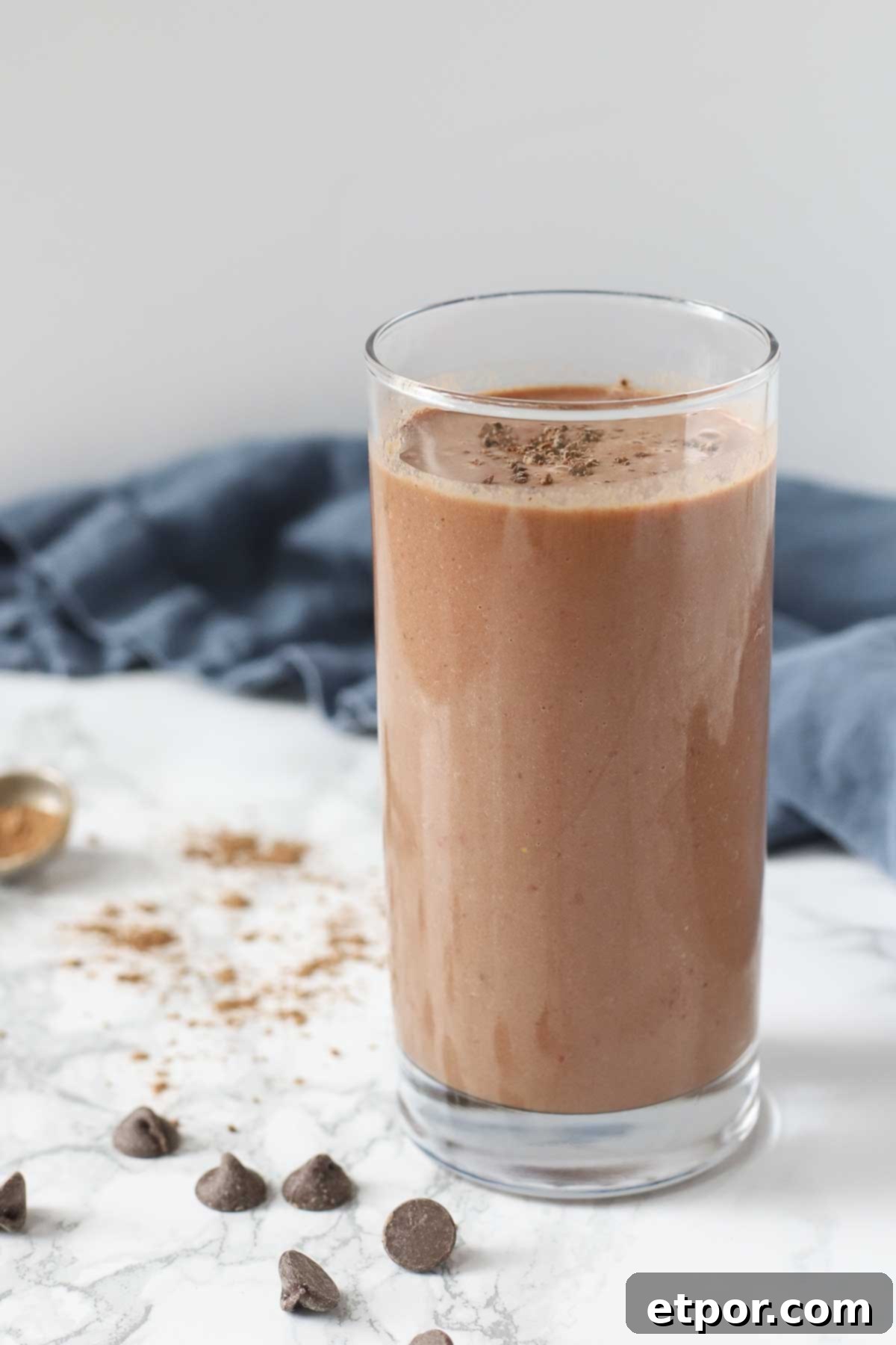 tall glass of a chocolate cauliflower smoothie on a marble countertop surrounded by chocolate chips, cocoa powder and a blue napkin