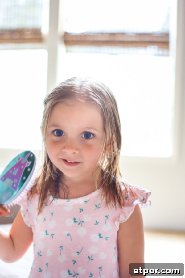 A little girl wearing a pink dress, smiling while brushing her wet hair with a detangling brush.