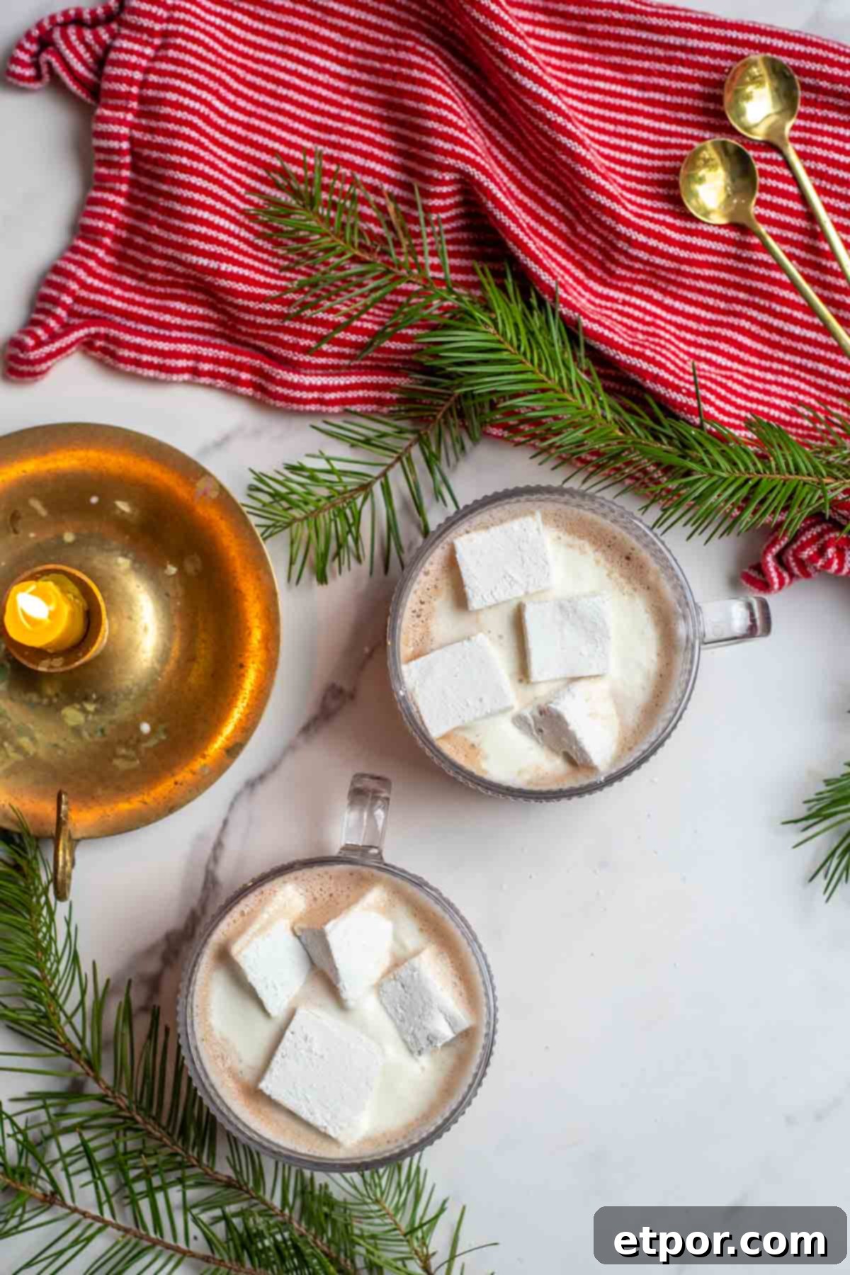 Two glass mugs of healthy hot chocolate topped with homemade marshmallows. Greenery, a candle, red towel with brass spoons surrounding the mugs.