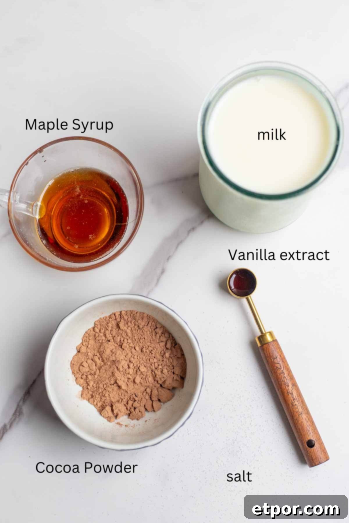 small bowls of cocoa powder and maple syrup, a glass of milk and a measuring spoon of vanilla on a countertop.