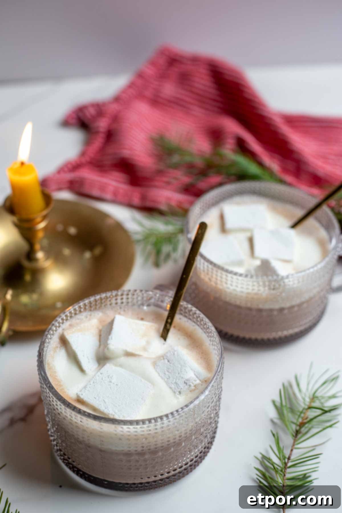 Two mugs of creamy hot chocolate topped with homemade marshmallows and whipped cream with brass spoons in each mug. a candle, towel and greenery are in the background.