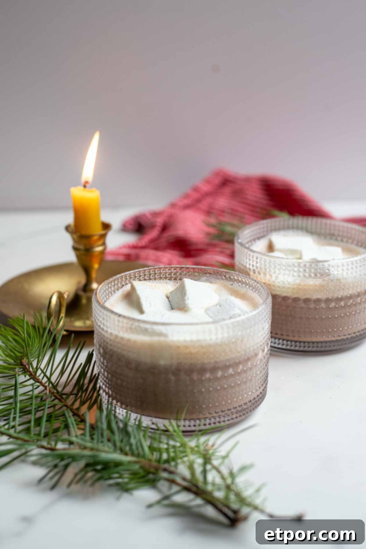 Two mugs of healthy hot chocolate topped with homemade marshmallows and whipped cream. The mugs are surrounded by greenery and a candle.