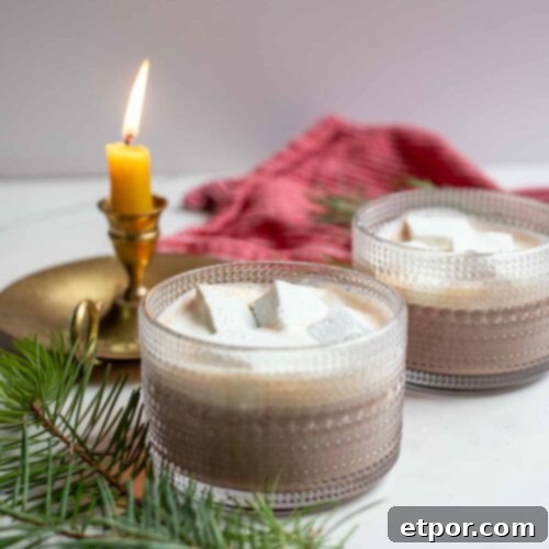 Two mugs of healthy hot chocolate topped with homemade marshmallows and whipped cream. The mugs are surrounded by greenery and a candle.