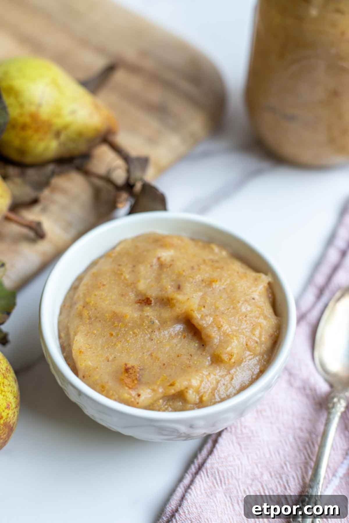 Homemade pear sauce in a bowl. A pink napkin with a spoon is to the right of the bowl, and cutting board with more pears are behind the bowl.