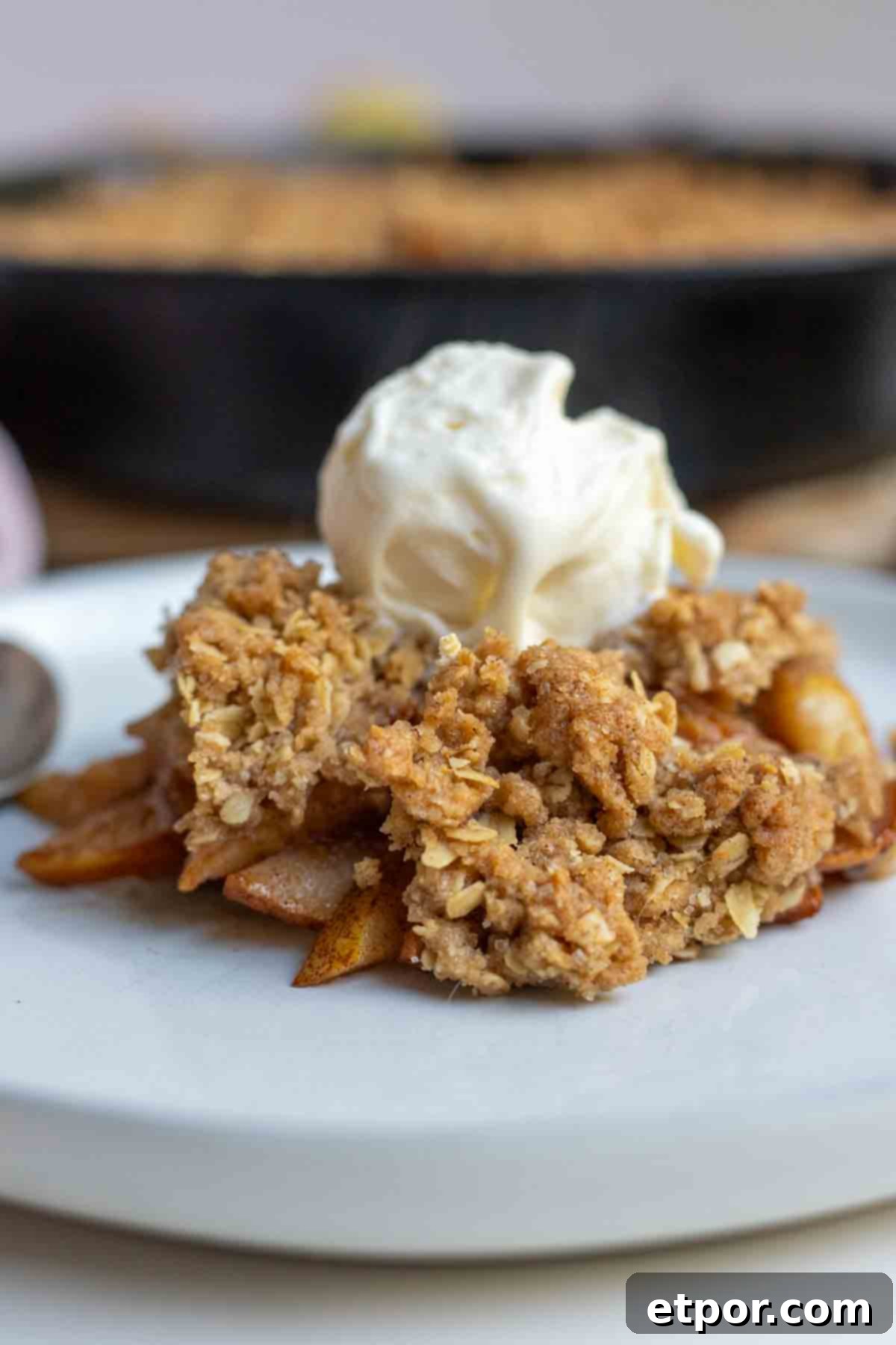 Pear crisp topped with a scoop of ice cream on top on a plate with a cast irons skillet of more crisp in the background.