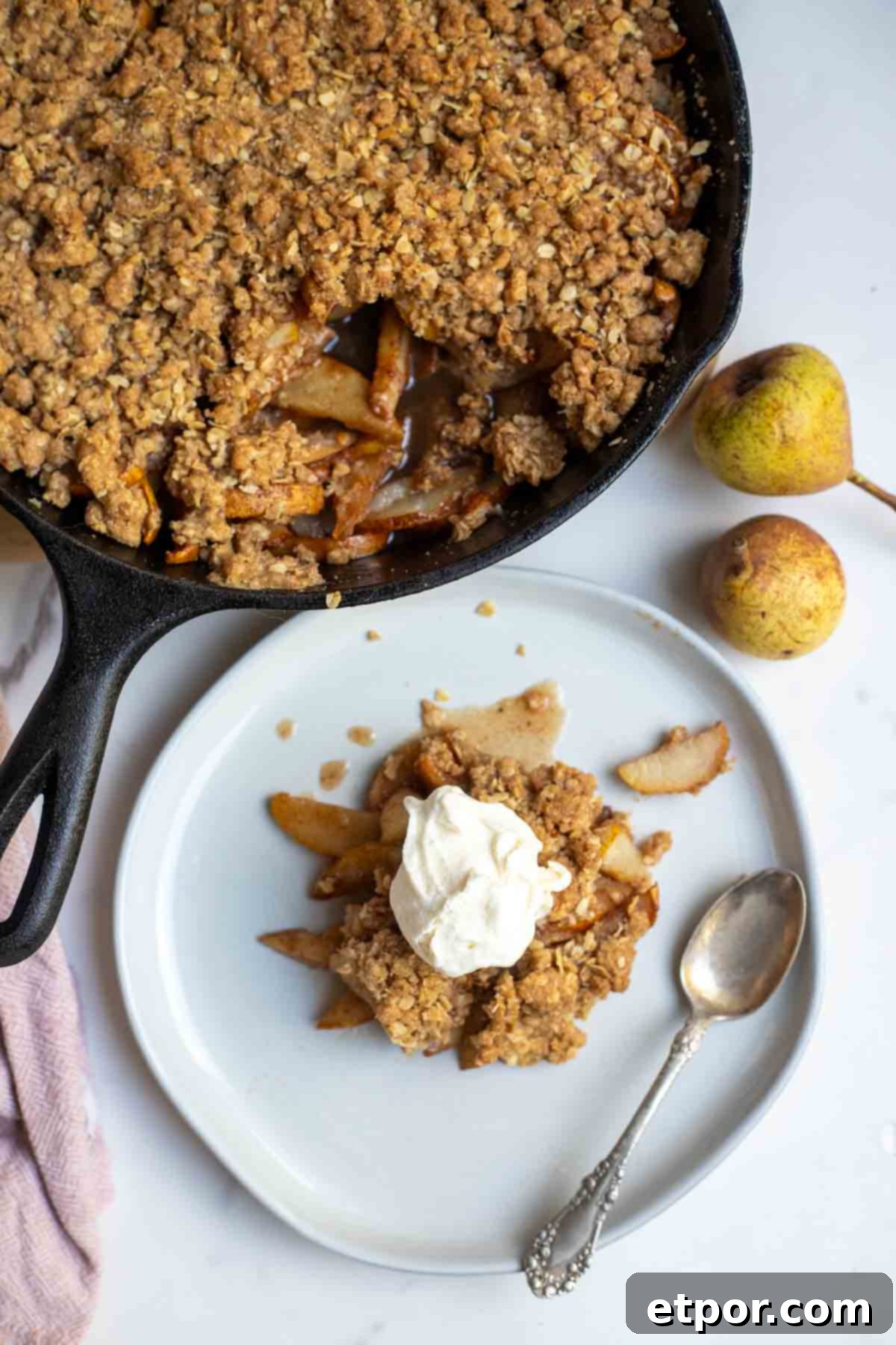 Overhead photo of pear crisp topped with ice cream on a plate with a spoon and a skillet of pear crisp in the background.