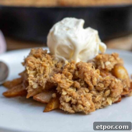 Pear crisp topped with a scoop of ice cream on top on a plate with a cast irons skillet of more crisp in the background.