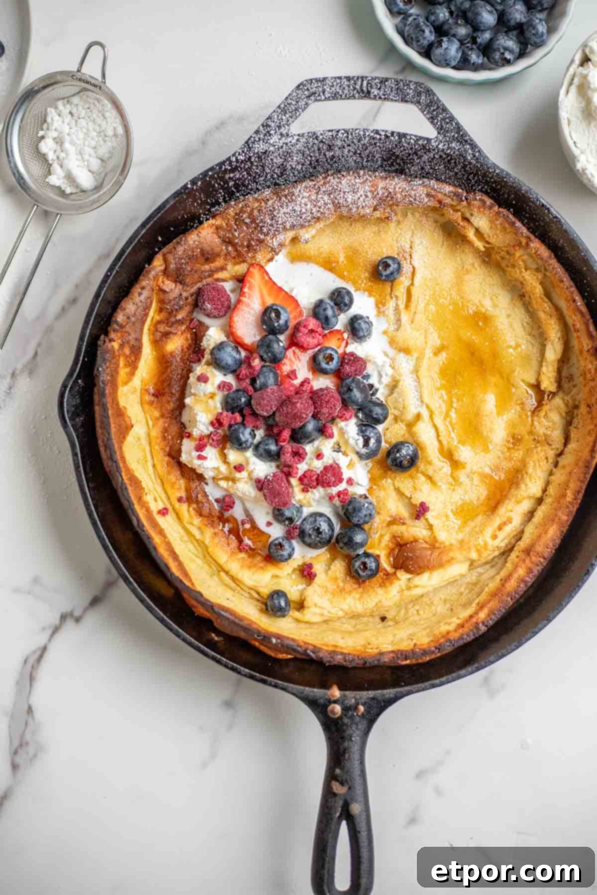 Dutch baby pancake topped with whipped cream, blueberries, strawberries, raspberries, and powdered sugar in a cast iron skillet. The skillet is surrounded by blueberries and a sifter of powdered sugar.