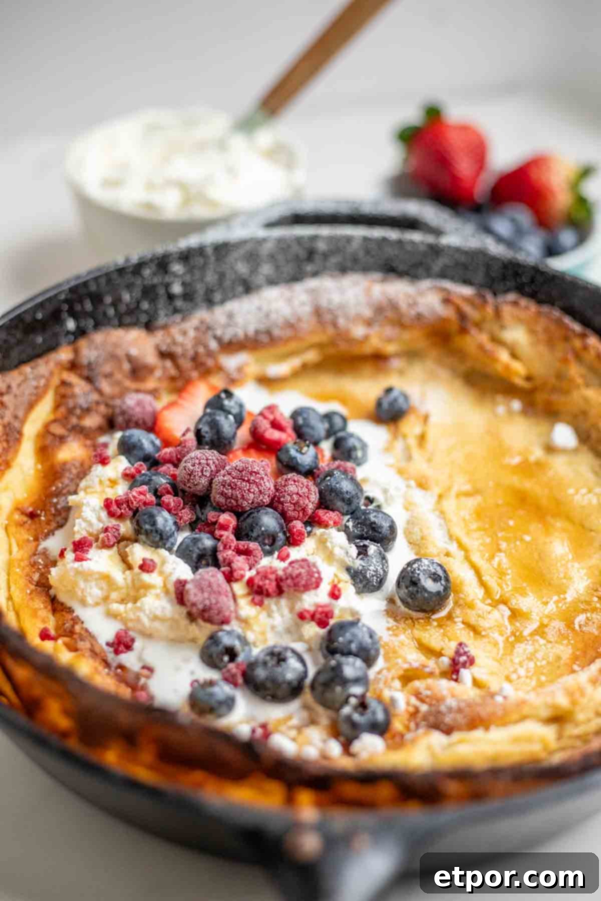 dutch baby pancake topped with whipped cream, berries, and powdered sugar. A Bowl of whipped cream and strawberries in the background.