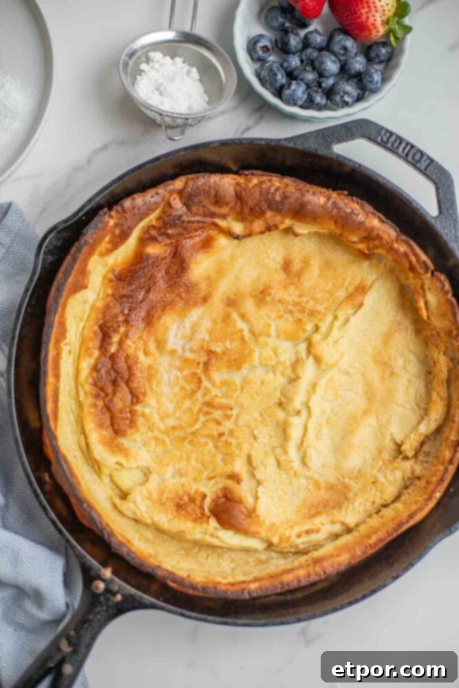 Baked dutch pancake in a skillet with blueberries and strawberries in a bowl, and powdered sugar in a stainer.