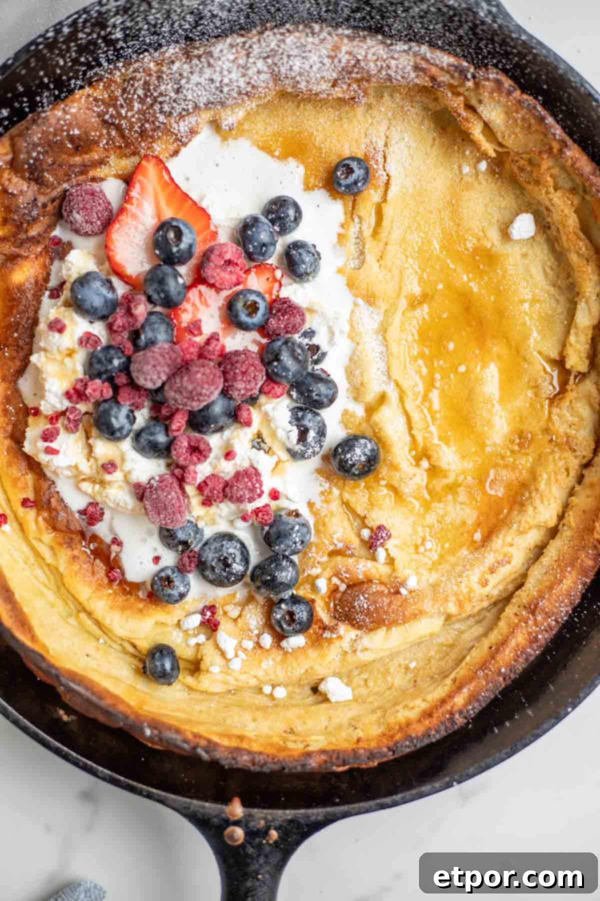 Overhead close up photo of a dutch baby pancake with whipped cream, berries, powdered sugar and maple syrup in a cast iron skillet.